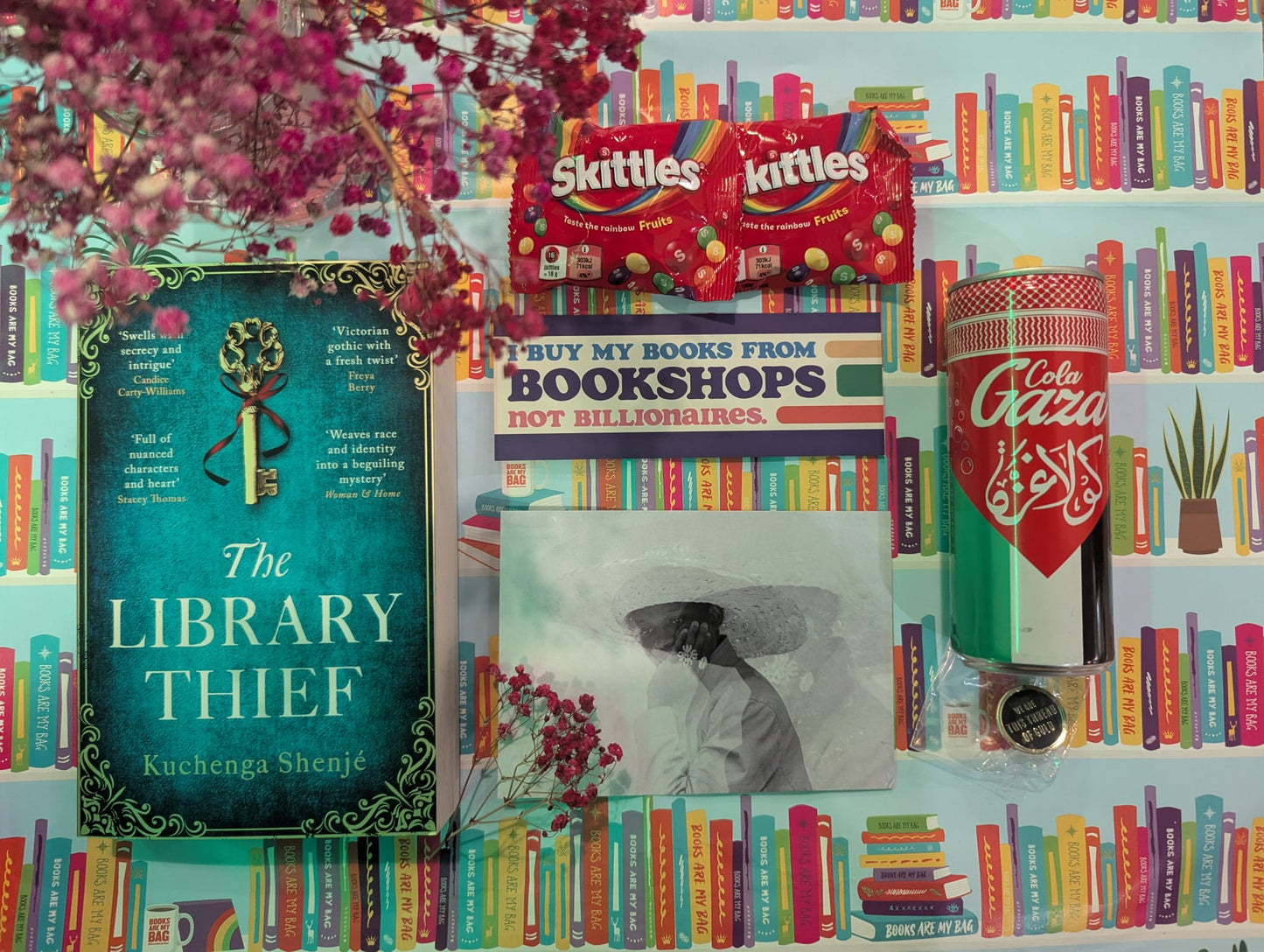Colourful book-themed background with 'The Library Thief' book, Skittles sweets, a gold pin badge and a Cola Gazza drink.